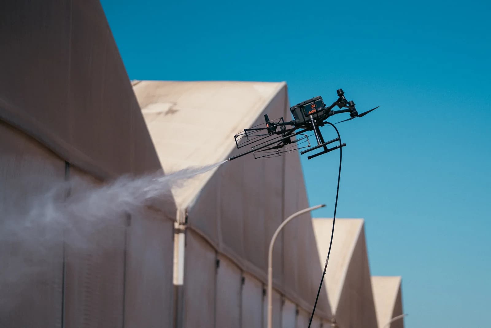 An AGS drone washing the façade of an industrial building with a pressurised water jet, its supply hose hanging beneath the airframe